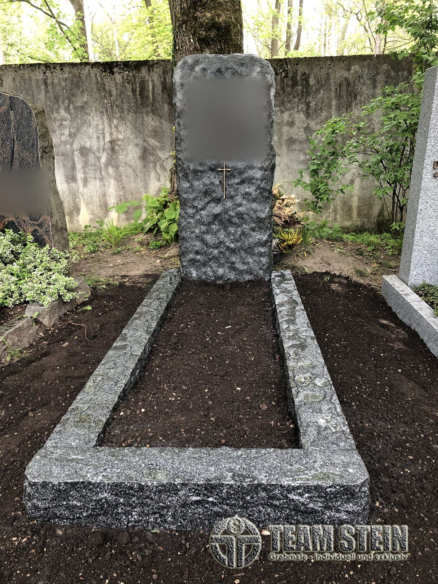 Rau behauener, dunkler Grabstein mit kleinem Kreuz und rauer Einfassung